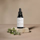 A black dropper bottle of Vitamin C Enriched Brightening Serum stands on a textured stone slab with small green and white flowers in the foreground, set against a warm beige background.