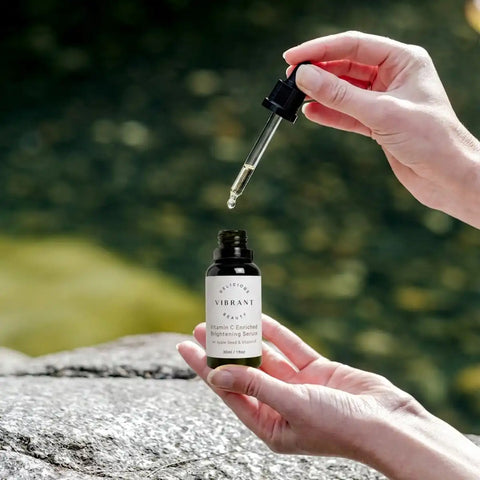 A person holds an open dropper bottle of serum outdoors, with the glass dropper hovering above the bottle. The background shows a blurred green natural setting with a stone surface in the foreground.