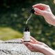 A person holds an open dropper bottle of serum outdoors, with the glass dropper hovering above the bottle. The background shows a blurred green natural setting with a stone surface in the foreground.