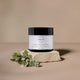 A jar of Vitamin Rich Day Cream sits on a textured stone slab with small white flowers, set against a solid beige background.