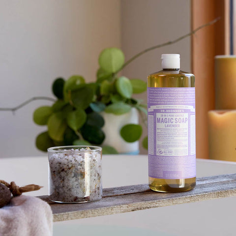 Bottle of Dr. Bronner's 18-in-1 Pure-Castile Magic Soap - Lavender on a surface with a candle & plant in the background.