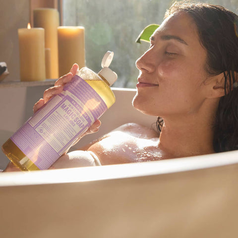 Woman in a bathtub holding a bottle of 
Dr. Bronner's 18-in-1 Pure-Castile Magic Soap - Lavender with candles in the background.