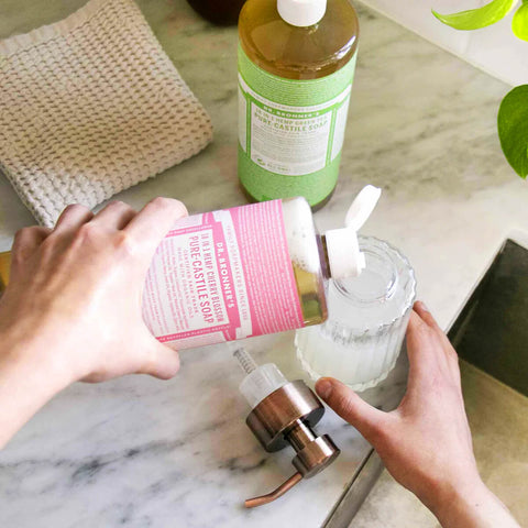Person using a pump bottle to dispense soap into a glass container on a marble countertop.