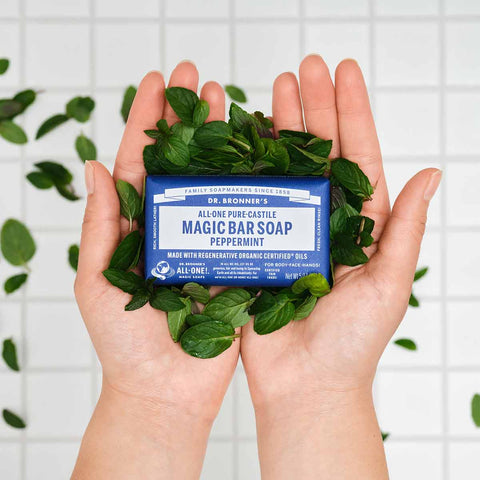 Bar of magic soap with a blue label held between two hands surrounded by mint leaves on a white tiled background
