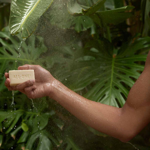 Hand holding a bar of peppermint magic soap under a shower with green leaves in the background
