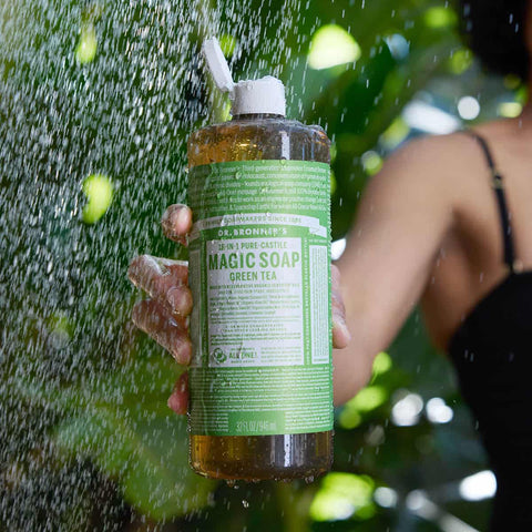 Person holding a bottle of Dr. Bronner's Magic Soap under a shower with greenery in the background
