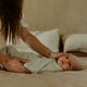 Baby wrapped in a muslin swaddle being held by an adult on a bed.