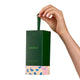 A hand holds a dark green Endota skincare box with a matching green handle against a clean white background. The box features the brand name 'endota' in green and a decorative bottom section with pink, blue, and beige patterns.