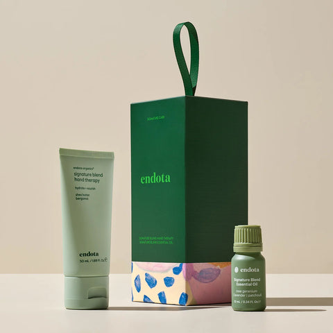 Endota gift set featuring a signature blended hand cream tube, a dark green gift box with a handle, and a small signature blended essential oil bottle, arranged on a light beige surface against a neutral beige background.
