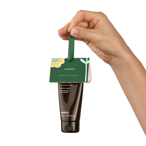 A hand holds a small Endota skincare tube with a green hanging tag against a plain white background. The tube is dark brown, and the tag is green with white text and a small yellow flower graphic.