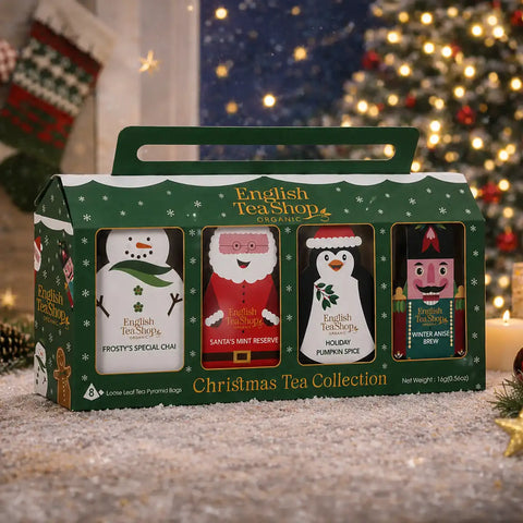 A boxed set of four English Tea Shop Christmas tea bags in a green gift bag with a handle, placed on a snowy surface in front of a blurred Christmas tree and holiday decor.