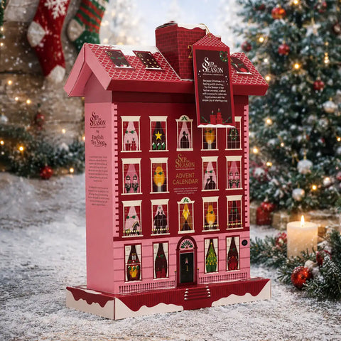 A pink and red house-shaped Advent Calendar displayed outdoors on a snowy surface with a decorated Christmas tree and candles in the background.