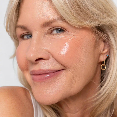 Close-up of a woman with blonde hair and a gold earring, wearing harmony shade of the carrot colour pot on her cheeks & lips against a white background.
