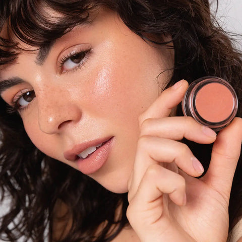 Woman holding up 'healthy' colour pot next her face with the product applied to her cheeks, lips & eye area. 