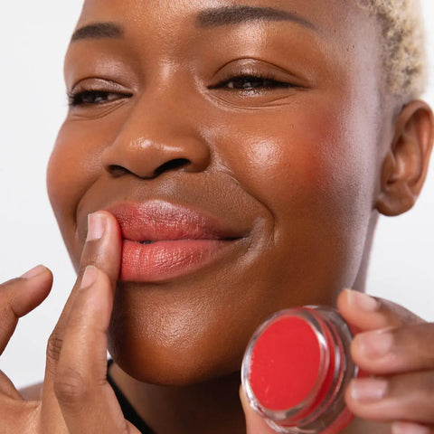 Woman applying red lip balm to her lips, with some on her cheeks, while holding pot near her face for a close-up view.