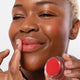 Woman applying red lip balm to her lips, with some on her cheeks, while holding pot near her face for a close-up view.