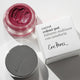 Magenta carrot colour pot in the shade 'holy' with an aluminium lid and white cardboard packaging next to it.