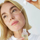 Woman applying a dropper of oil to her face with a bottle of face nectar in her other hand.
