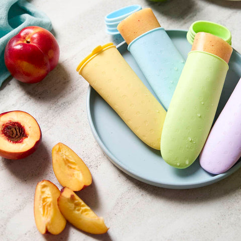 Colourful popsicle moulds filled with frozen fruit puree on a plate with peaches and peach slices on a light surface.