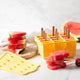 Yellow silicone popsicle tray - set of 12 with watermelon popsicles with some stacked next to the tray with the lid to the side and some strawberries & sliced watermelon.