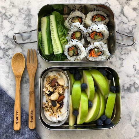 Two stainless steel bento boxes packed with sushi and apple, placed beside bamboo cutlery on a marble surface.