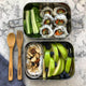 Two stainless steel bento boxes packed with sushi and apple, placed beside bamboo cutlery on a marble surface.