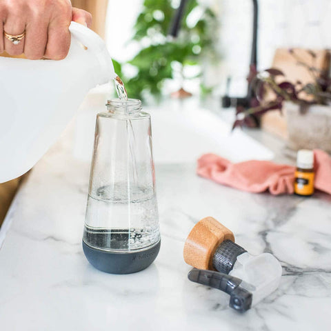 Glass bottle with lid off while a person pours in water, styled on a marble counter with cleaning cloth & essential oils in the background.
