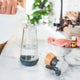 Glass bottle with lid off while a person pours in water, styled on a marble counter with cleaning cloth & essential oils in the background.