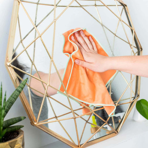 Hand holding an orange cloth against a geometric gold framed mirror with plants in the background.