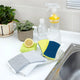 A set of cleaning supplies arranged on a kitchen counter near a sink, including two scrubber sponges, a lemon half, a clear spray bottle, and a small potted plant.