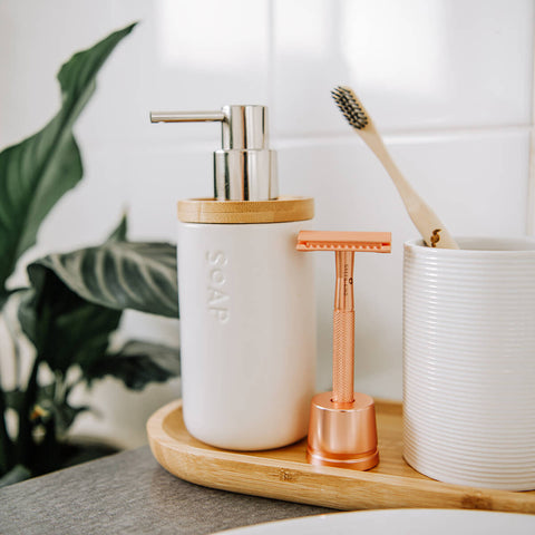 Bathroom setting with a soap dispenser, razor, and toothbrush on a wooden tray.