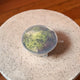 A silver bowl with a transparent silicone lid, shown filled with green lettuce and placed on a round speckled stone surface.
