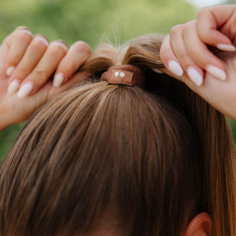 Close-up of a person adjusting a Kooshoo Plastic-free hair tie on their hair with a blurred green background.