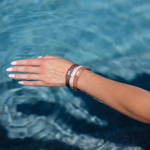 A hand wearing Kooshoo plastic-free hair ties on the wrist, touching clear blue water.