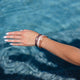 A hand wearing Kooshoo plastic-free hair ties on the wrist, touching clear blue water.