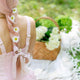 Person with braided pink/blonde hair featuring daisies, and Kooshoo's Satin Bow. Wearing a pink dress, sitting on a blanket with a basket of flowers in a garden setting.