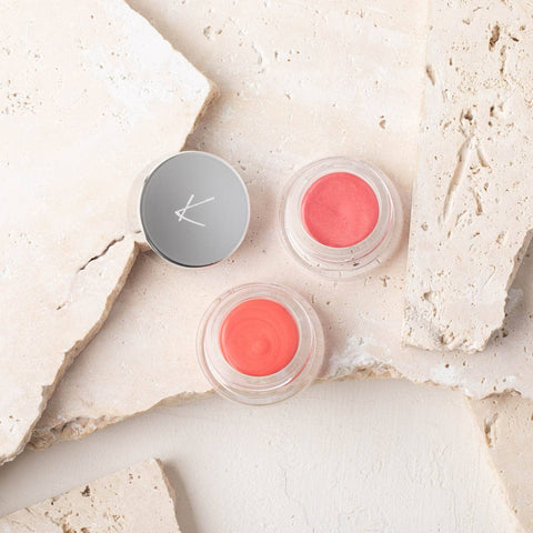 Two small containers of pink & coral-coloured cheek & lip cream on a textured beige surface.