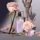 A clear perfume bottle stands on a grey surface surrounded by pink roses, vanilla beans, lavender sprigs, and soft pink petals.
