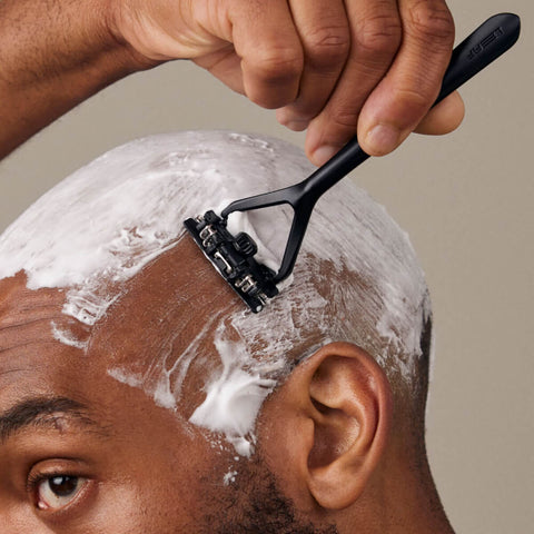 Person shaving their head with a razor and shaving cream on a neutral background