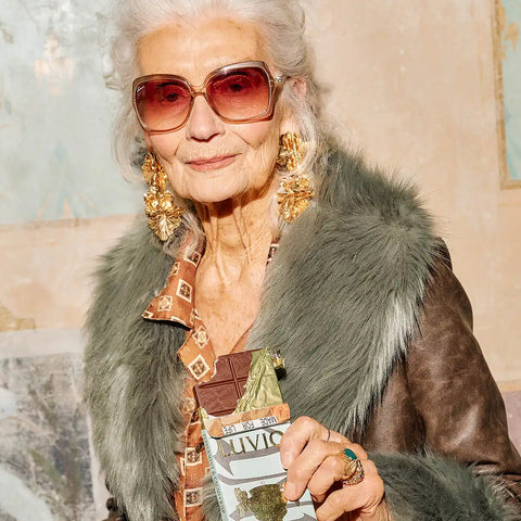 Elderly woman wearing sunglasses and a fur coat, holding an open Luvio peppermint bar with a bite taken out.