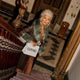 Posh woman with a Luvio bag and chocolate bar peeking out. Dressed to the nines, standing on a grand staircase, with purple carpet, with a sculpture in the background.