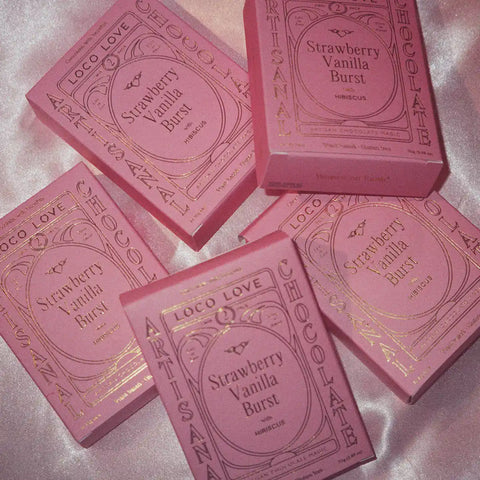 Pink packaging of Strawberry Vanilla Burst chocolate bars on a light pink silk background.