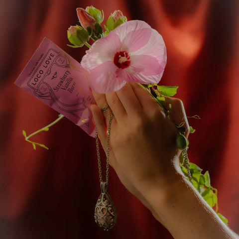 Hand holding a pink hibiscus flower and a small pink packet with 'LOCO LOVE' branding against a dark background.