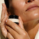 A close-up lifestyle shot of a woman holding a small black jar of MUKTI Eye Creme against her face.