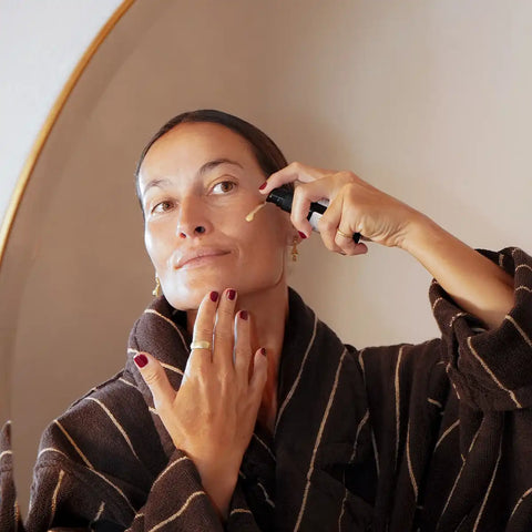 A woman wearing a brown striped bathrobe applies a serum from a pump bottle to her face.