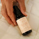 A hand holds a small black pump bottle of MUKTI Bioactive Renewal Night Serum against a soft white quilted fabric background.