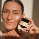 A woman holds a small jar of Queen of the Night Creme next to her cheek where a dollop of the cream is applied, in warm natural light.