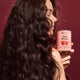 A woman with long, dark, wavy hair holds a pink tin labeled 'Hair Ritual' against a deep burgundy background.
