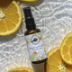 A small amber spray bottle labeled 'Perfect Potions Happy and calm aromatic mist' placed on a glossy white surface with sliced orange slices around it.