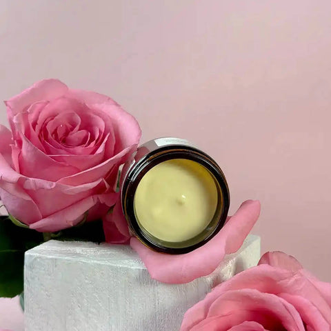 An open round cosmetic jar filled with pale yellow replenish ultra rich moisture cream sits on pink rose petals beside a large pink rose, set against a soft pink background.